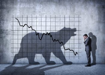 A businessman covers his eyes as he can't watch a descending stock chart and an ominous shadow of a bear that is cast on the wall above him.