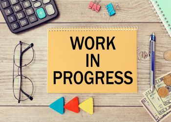 Blank notepad with text Work in progress on wooden surface, calculator, glasses, money