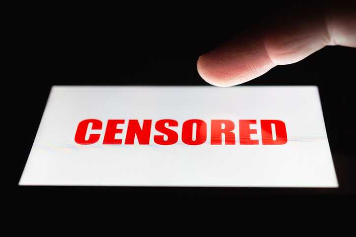 A smartphone lying on a table in the dark, displaying "censored" text. The finger above touch screen. The concept of censorship on popular social networks. Restricted access to internet. Shallow DOF