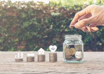 Savings money for family life concepts. Hand holding coin on a full money in glass jar and family member, car, house, healthy on coins. Depicts saving for wealth and life. fundraising concept.