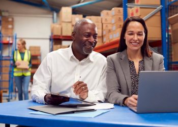 Male And Female Team Leaders Working On Laptop In Warehouse With Workers In Background