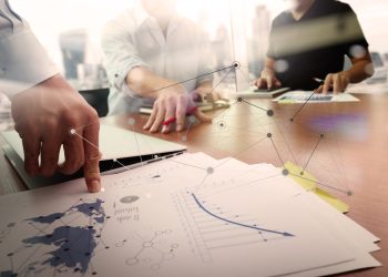 business documents on office table with smart phone and laptop computer and graph financial with social network diagram and two colleagues discussing data in the background